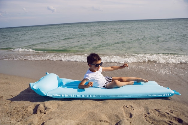 Boy child in a white T-shirt and striped swimming trunks and sunglasses is lying on a blue inflatable mattress on the beach the sailor