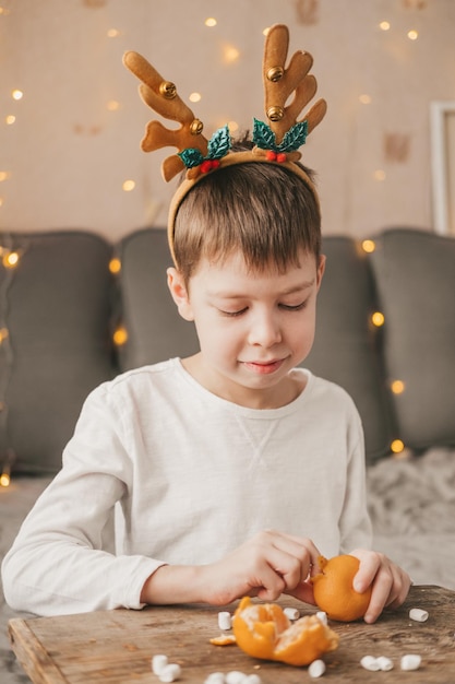 Boy in Christmas antlers, sits and peels a tangerine on the background of a yellow garland. Boy in a white t-shirt at home, peels a tangerine on a wooden table with marshmallows