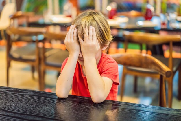 The boy closes his eyes with his hands while sitting at the table