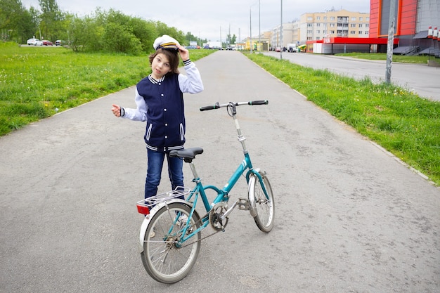 Boy in college jacket and cap standing with bicycle on asphalt road on summer street with greenery and houses.