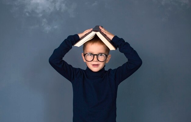 Photo boy covering his head with book on blue studio background portrait of funny smart schoolboy looks and smiles at camera concept of education
