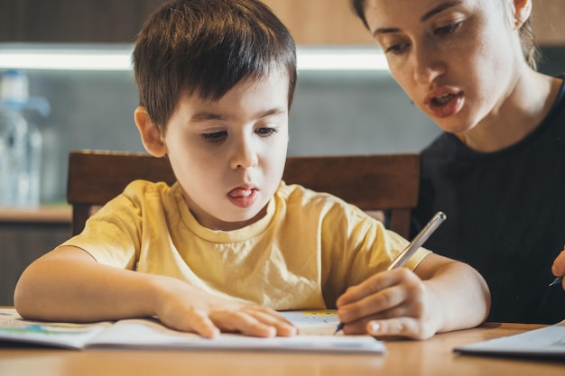 Boy doing his homework with his mother sitting at desk, at home, he is writing on copybook. Child development.