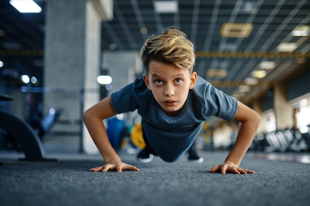 Мальчик делает упражнения в тренажерном зале, вид спереди. Юноша на тренировках в спортклубе, здоровье и здоровый образ жизни, школьник на тренировке, спортивная молодежь
