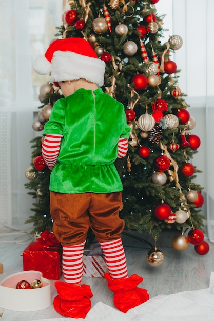 A boy dressed as a gnome and wearing a New Year's hat decorates a Christmas tree at home. Christmas.