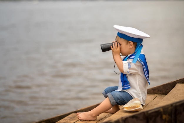 Photo a boy dressed as a sailor sits on the shore and looks through binoculars