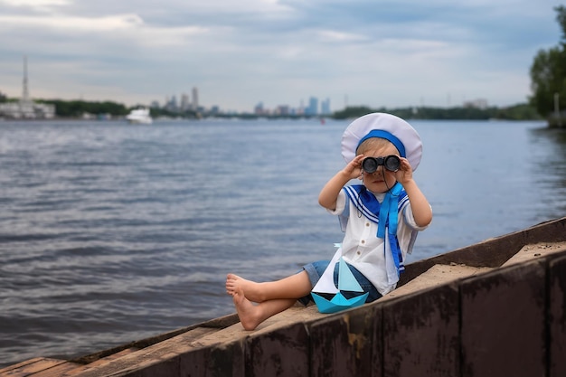 A boy dressed as a sailor sits on the shore with a paper boat and looks through binoculars