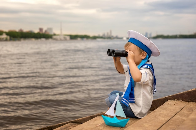 A boy dressed as a sailor sits on the shore with a paper boat and looks through binoculars