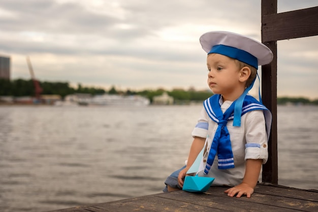 A boy dressed as a sailor sits on the shore with a paper boat