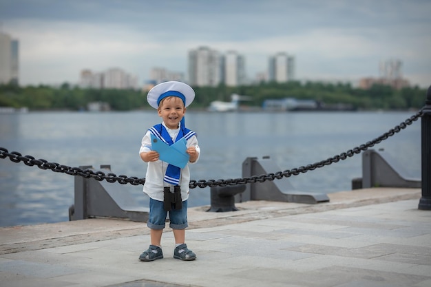 A boy dressed as a sailor with binoculars and a boat stands on the shore and laughs