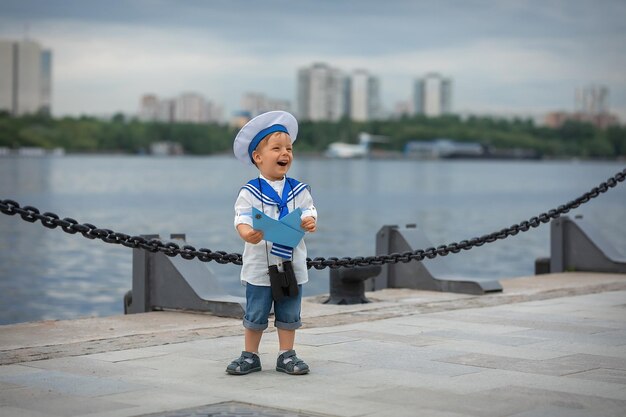 A boy dressed as a sailor with binoculars and a boat stands on the shore and laughs