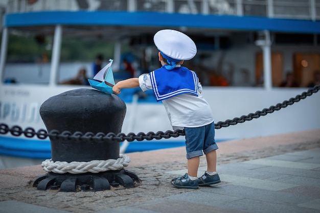 A boy dressed as a sailor with binoculars and a boat stands on the shore and looks at the ships