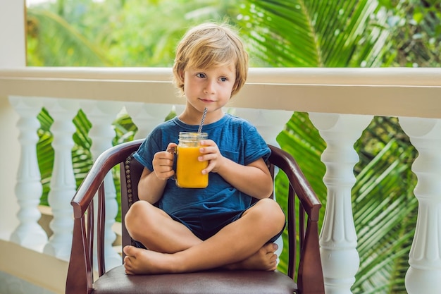 Мальчик пьет сочный коктейль из манго в стеклянной банке каменщика с полосатой красной соломой на старом деревянном фоне. Концепция здорового образа жизни, копией пространства
