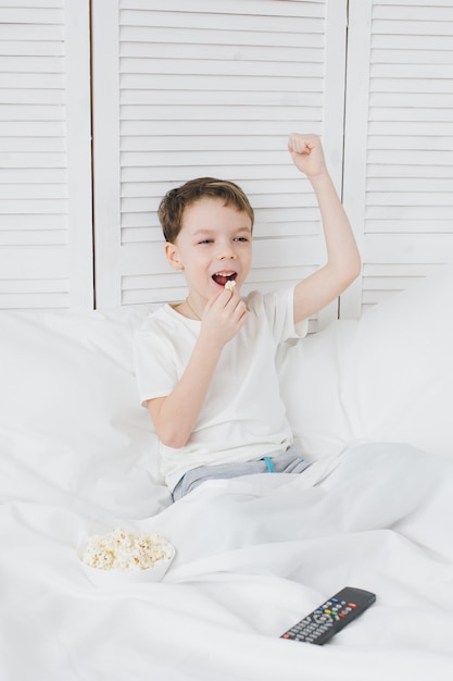 Мальчик ест попкорн, сидя в кровати и смотреть телевизор