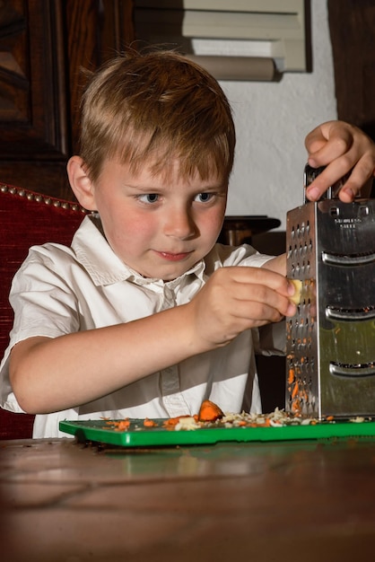 8歳の男の子がおろし金でサラダをこすり、本物の写真をカットする準備をします