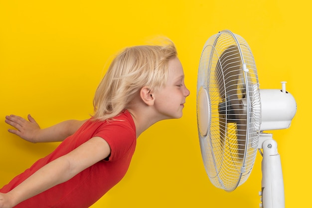 Boy enjoying cool air. Child and fan on yellow surface. Room temperature regulation.