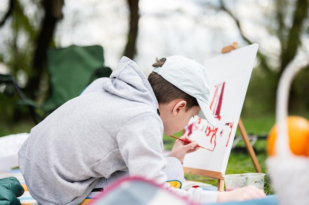 ピクニック毛布で屋外を楽しんだり、庭の春の公園のリラクゼーションでキャンバスに絵を描いたりする少年