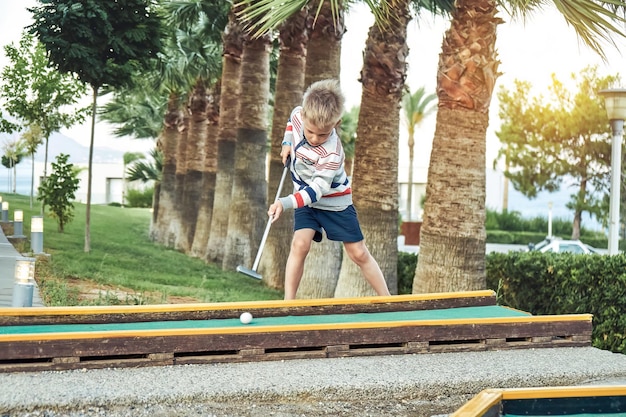 Boy enthusiastically plays mini golf on summer evening on the territory of the hotel