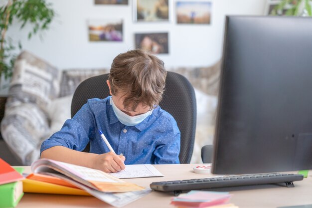 Boy in face mask using computer, doing homework during coronavirus quarantine