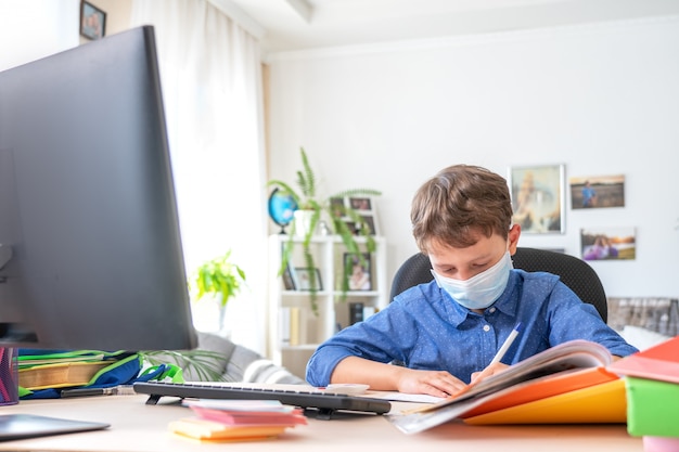 Boy in face mask using computer, doing homework during coronavirus quarantine