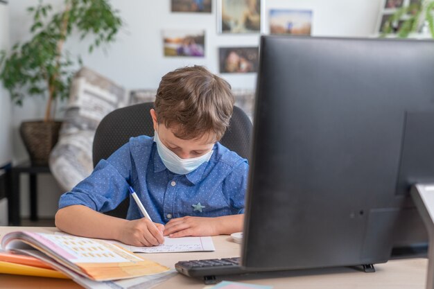 boy in face mask using computer, doing homework during coronavirus quarantine