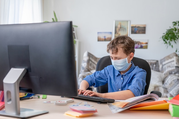 Boy in face mask using computer, doing homework during coronavirus quarantine