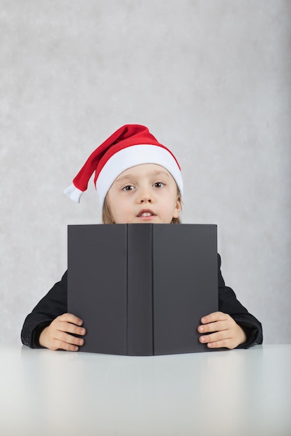 Boy of four years in Santa's hat and black classical jacket. Closeup  Boy of four years in Santa's hat and black book sits at the table. Closeup