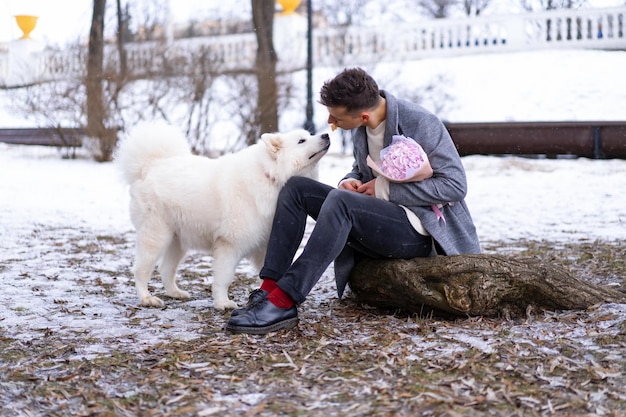 Ragazzo amico con un mazzo di fiori rosa ortensia che aspetta la sua ragazza e cammina e gioca con un cane. all'aperto mentre cade la neve. concetto di san valentino, proposta di matrimonio. l'uomo va