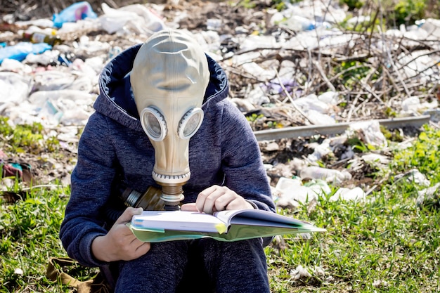 Boy in  gas mask reads  book on the background of  garbage. Ecological catastrophe