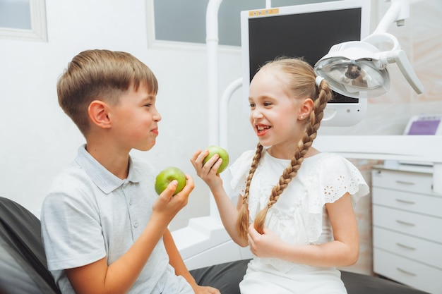 A boy and a girl are eating apples in the dentist\'s chair the\
concept of healthy teeth children eat a green apple