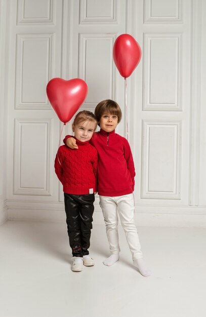 A boy and a girl hug and hold red balloons in the shape of a heart on white