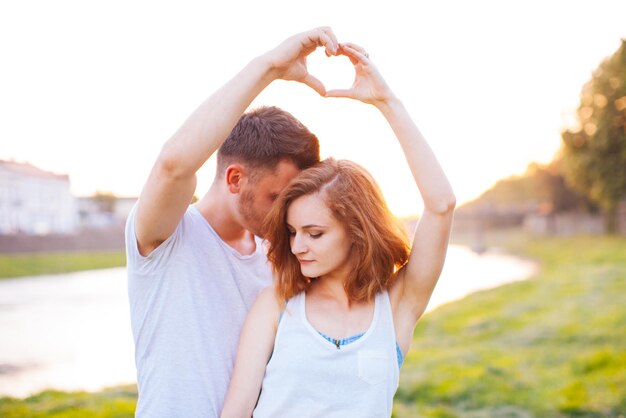 The boy and the girl make hands in the form of a heart against the background of the sunset