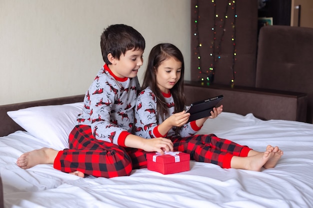 A boy and a girl in red and gray pajamas are sitting at home on the bed