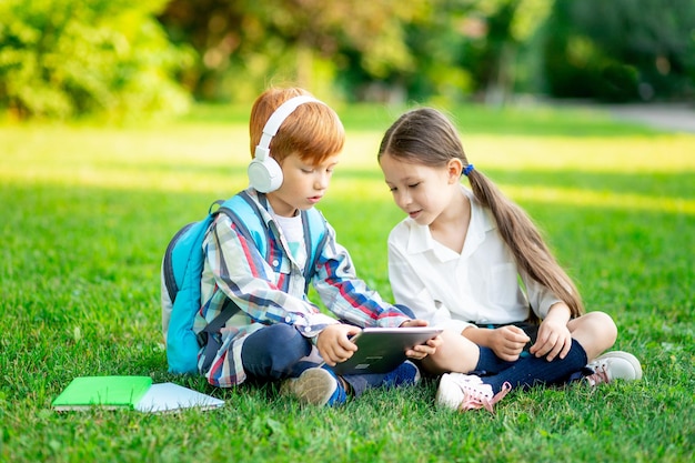 バックパックを持った男の子と女の子が芝生の上に座って本を読んでいるし、ヘッドホンでタブレットを読んでいるコンセプトは新学期の兄と妹が路上で宿題をしている