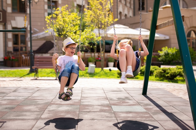 Boy in the glasses and hat and blonde girl in the dress having fun on a swing together in beautiful summer garden.