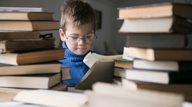 Boy in glasses reading a book with a stack of books next to him.