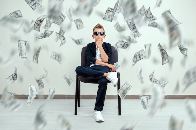  boy  glasses sits on a chair and holds a bundle of dollars in his hands. Knowing how to make money.