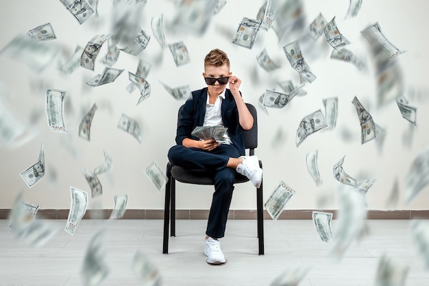 boy glasses sits on a chair and holds a bundle of dollars in his hands. Knowing how to make money.