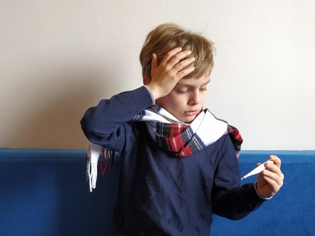 the boy got sick his head hurts viral disease is progressing the childs neck is wrapped in a warm scarf the kid looks dejectedly at the thermometer holds his hand on his forehead and suffers