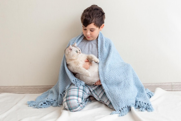 A boy in gray pajamas sits on the floor on a knitted blue blanket with a beautiful white cat