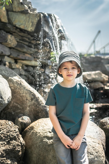 a boy in a gray panama hat sits on a large rock near the water