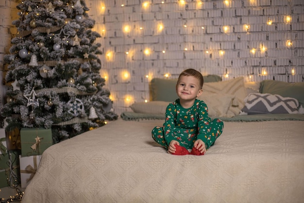 Foto un ragazzo in pigiama verde è seduto sul letto e aspetta che arrivi il nuovo anno le luci lampeggiano da dietro e c'è un elegante albero di natale