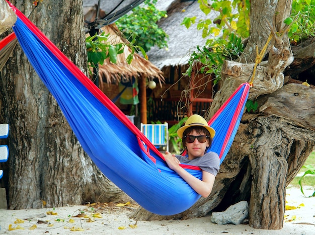 boy in a hat and sunglasses in a blue hammock on the beach