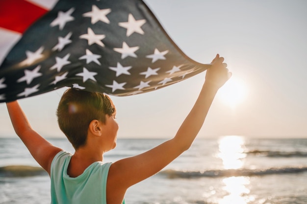 Photo boy holding american flag at beach during sunset