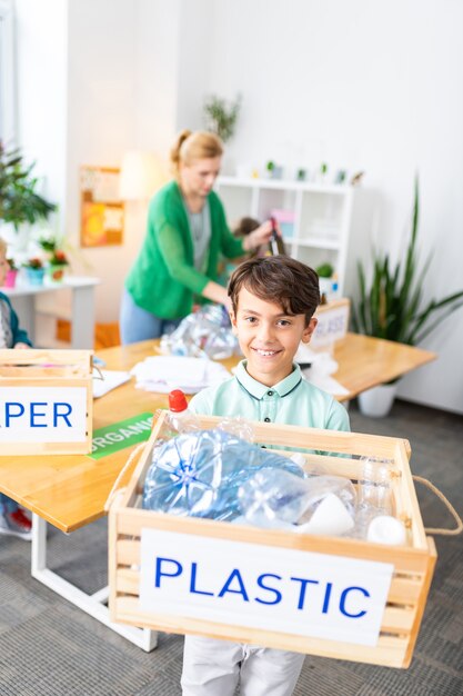 Boy holding box. Beaming handsome dark-haired boy holding box with plastic after waste sorting at school