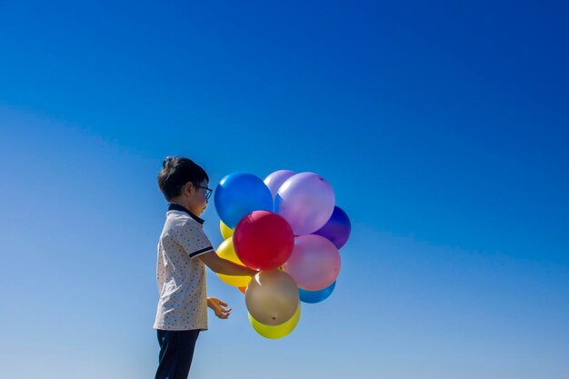 Foto ragazzo con palloncini multicolori in piedi contro un cielo blu limpido