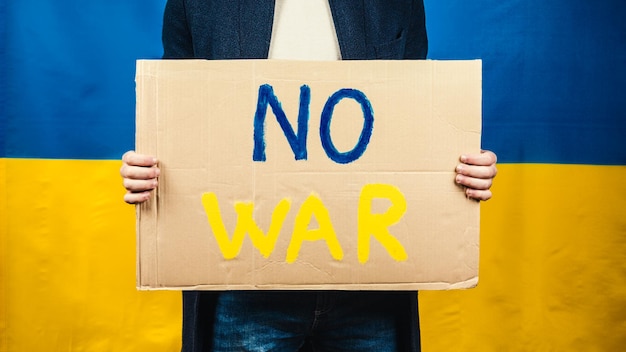 Boy holds in hand cardboard with no war text on ukrainian flag background