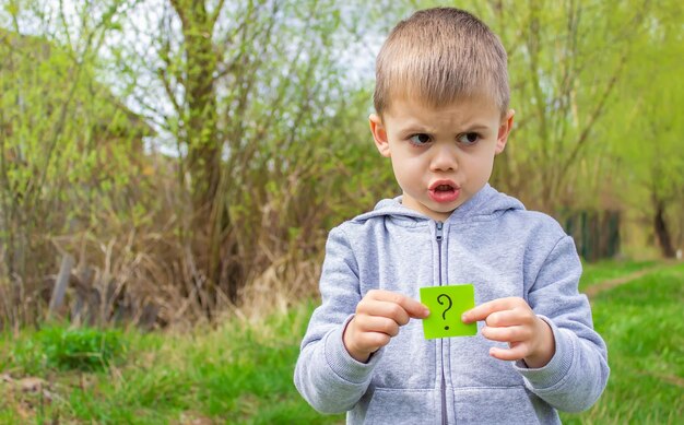 The boy holds stickers with a question mark on the background of the park selective focus