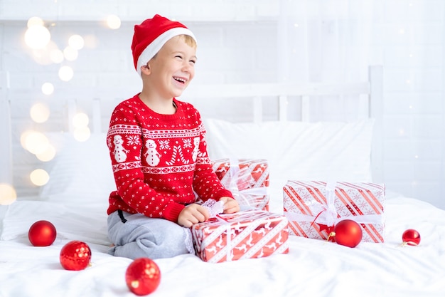 Фото Мальчик в шапке санты и красном свитере сидит с новогодними подарками на кровати радуется и смотрит на подарки