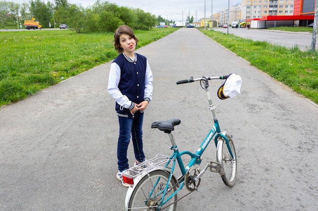 Фото Мальчик в куртке и кепке колледжа, стоящий с велосипедом на асфальтовой дороге на летней улице с зеленью и домами.