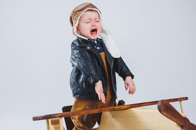 The boy is crying in a leather jacket and a pilot's hat a wooden plane brown suitcases Children's wooden toys Baby emotions